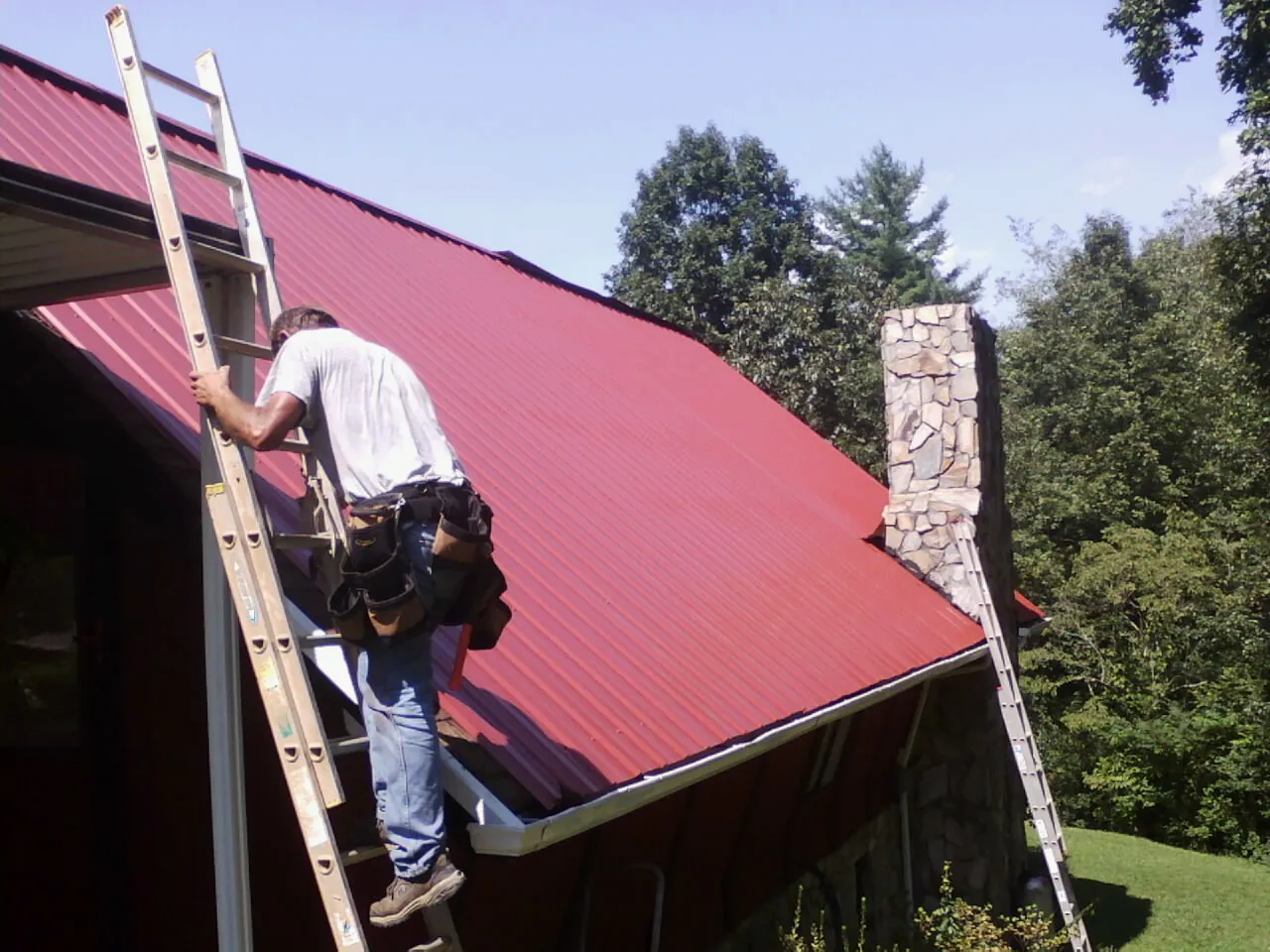 Roofer on a ladder beside a red metal roof and stone chimney — Soffit & Fascia Repair by Roofing Brothers in Catawba